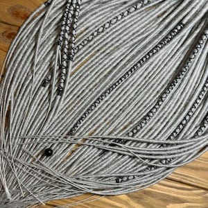 May include: Close-up of silver-gray dreadlocks with black thread accents. The dreadlocks are arranged in a layered pattern, with a few strands extending outwards. A small black bead is visible near the bottom. The background is a wooden surface.