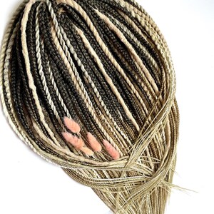 Può includere: Un fascio di dreadlock sintetici in varie tonalità di marrone, beige e biondo, con alcuni accenti decorativi rosa. I dreadlock sono disposti in un motivo circolare, con ciocche sciolte nella parte inferiore.