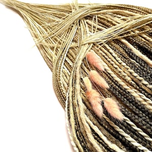 Può includere: Primo piano di un fascio di dreadlock sintetici in varie tonalità di marrone, beige e bianco. I dreadlock sono adornati da diversi rametti di fiori secchi soffici, rosa chiaro. Lo sfondo è bianco.