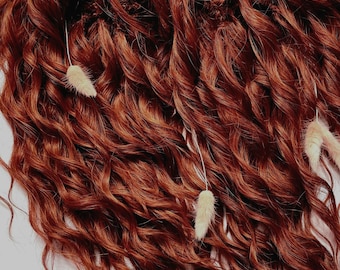 May include: Close-up of auburn curly hair extensions. The extensions have a wavy texture and are decorated with small, dried, beige floral accents. The hair is arranged in a loose, flowing pattern. The hair is a rich, warm shade.