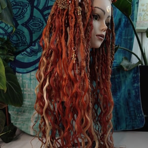 May include: A mannequin head featuring long, red and blonde dreadlocks, adorned with gold charms and white beads. The mannequin has a gold crescent moon on its forehead. The backdrop is a blue patterned fabric.