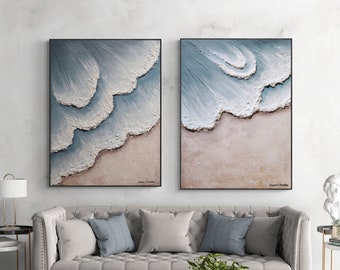 May include: Two framed abstract paintings of ocean waves crashing on a sandy beach. The paintings are in shades of blue, white, and beige. The paintings are hanging on a white wall above a gray sofa with pillows.