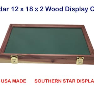 May include: A cedar wood display case, 12 x 18 x 2 inches, with a dark green interior and a wooden frame. The case has two gold-colored latches and is designed for displaying collectibles. The text "Cedar 12 x 18 x 2 Wood Display Case" is visible.