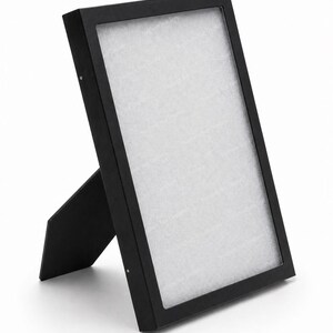 Puede incluir: Un marco de fotos rectangular negro con un respaldo blanco, sostenido por un soporte. El marco está en ángulo, destacando sus líneas limpias y su diseño minimalista. El marco está hecho de un material negro mate.