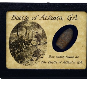 May include: A black display case with a beige background featuring a black and white illustration of soldiers in a wooded area. The text "Battle of Atlanta, GA." is printed above the illustration. A bullet is displayed in a circular cutout on the right side of the case. The text "Shot bullet found at The Battle of Atlanta, GA." is printed below the bullet.