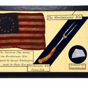Könnte beinhalten: Eine gerahmte Vitrine mit einer Nachbildung der amerikanischen Flagge mit 13 Sternen, einer Gabel aus der Zeit des Amerikanischen Unabhängigkeitskriegs mit einem Knochengriff und einem Stück zerbrochener Keramik aus der Zeit des Amerikanischen Unabhängigkeitskriegs. Der Text "The Revolutionary War" befindet sich oben auf der Vitrine.