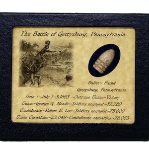 Könnte beinhalten: Gerahmte Präsentation mit einer Kugel, die in der Schlacht von Gettysburg, Pennsylvania, gefunden wurde. Die Präsentation enthält eine Illustration der Schlacht, Text mit Datum, Ergebnis und Opferzahlen. Die Kugel ist in einem dunkelblauen Oval eingeschlossen.