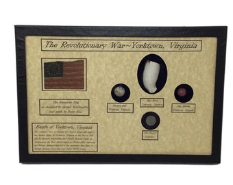 May include: A framed display with a brown background and black frame. The display features a replica of the American flag, a pipe bowl, a musket ball, a clay marble, and a flat button. The text "The Revolutionary War~Yorktown, Virginia" is at the top of the display. The text "Battle of Yorktown, Virginia" is at the bottom of the display.