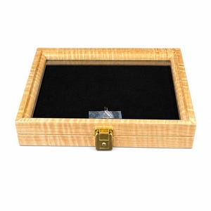 May include: A rectangular wooden display case with a glass top and a black felt interior. The case has a light wood finish, a gold-colored latch, and a small object inside. Ideal for displaying collectibles.