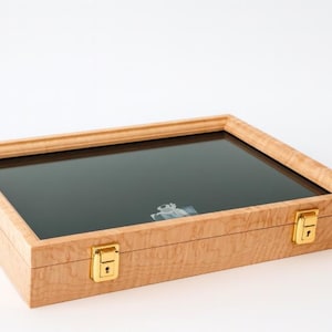 Op de afbeelding: Een houten vitrine met een glazen bovenkant en goudkleurige grendels. De behuizing is gemaakt van lichtgekleurd hout met een zichtbaar nerfpatroon. De glazen bovenkant is donker en de behuizing lijkt leeg.