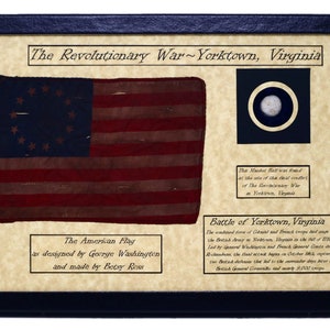 Op de afbeelding: Een ingelijste weergave met een replica van de Amerikaanse vlag met 13 sterren in een cirkel, een musketkogel en tekst die de Slag om Yorktown, Virginia, beschrijft. De tekst luidt "The Revolutionary War - Yorktown, Virginia", "The American Flag as designed by George Washington and made by Betsy Ross" en "Battle of Yorktown, Virginia".