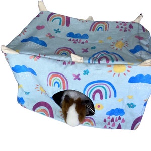 May include: A blue and white hanging pet bed with a rainbow and sun pattern. The bed has a hole for a pet to enter and a guinea pig is peeking out of the hole.