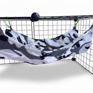 May include: A soft, fleece-like fabric hammock featuring a grey, white, and black camouflage pattern. The hammock is suspended from a black wire grid structure using four bright green metal carabiners attached to the corners of the fabric.