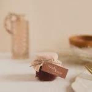 Peut inclure: Un petit pot rempli d'une confiture rouge foncé, recouvert d'un tissu et attaché avec de la ficelle. Une petite étiquette marron avec le mot "Yara" est attachée. Le pot est posé sur une surface blanche.