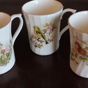 May include: Three white ceramic mugs with gold rims and handles. Each mug features a different bird illustration with floral accents. The birds are yellow, brown, and orange. The mugs have a ribbed design.