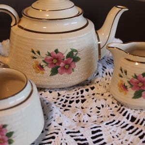 Peut inclure: Un ensemble à thé vintage de couleur crème, comprenant une théière, un pot à crème et un sucrier. Chaque pièce est ornée de motifs floraux roses et jaunes et de bordures dorées. L'ensemble est présenté sur un napperon en dentelle blanche.