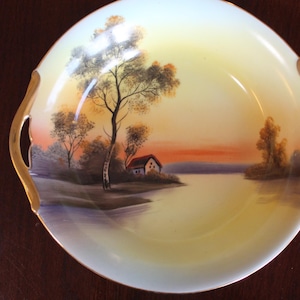 Peut inclure: Un plat de service rond vintage avec une scène de paysage peinte à la main. Le plat présente une vue paisible au bord du lac avec une petite maison, des arbres et un coucher de soleil. Il a des poignées dorées et un intérieur jaune.