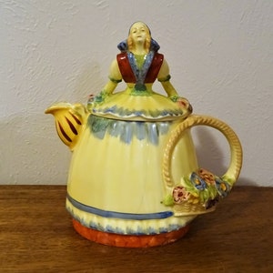 May include: A yellow ceramic teapot with a hand-painted design of a woman in a blue and red dress. The teapot has a wicker handle and a floral design on the side.