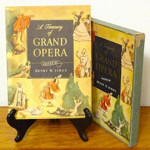 Puede incluir: Se muestran dos libros antiguos titulados "A Treasury of Grand Opera". Las portadas presentan ilustraciones de escenas de ópera en tonos naranja, amarillo y azul. El libro de la izquierda está apoyado en un soporte negro, mostrando la portada.