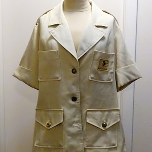 Puede incluir: Chaqueta beige claro de manga corta con cuello con muescas, dos bolsillos en el pecho y dos bolsillos inferiores. La chaqueta tiene una parte delantera abotonada y un pequeño parche bordado en el pecho. Las mangas están dobladas.