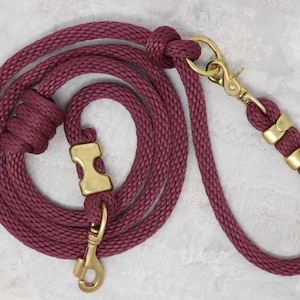May include: A maroon dog leash with brass hardware, including a clip, a ring, and a stopper. The leash is made of braided rope.