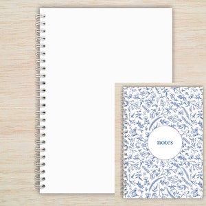 May include: Two spiral bound notebooks. The larger notebook has a white cover. The smaller notebook has a white cover with a blue floral pattern and the word "notes" in a circle.