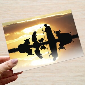 May include: A Christmas card with a silhouette of the Nativity scene set against a golden sunset. The card shows Mary, Joseph, baby Jesus, animals, and a shepherd with a staff. The image is held by a hand.