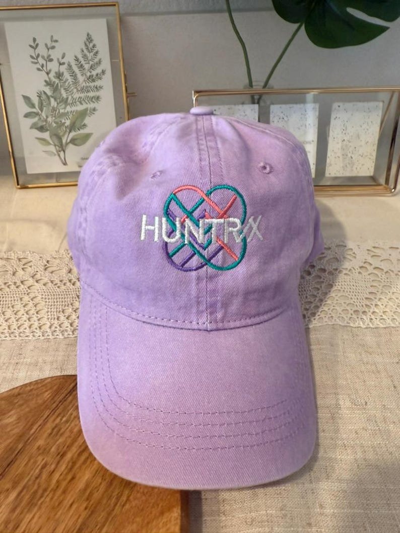 Puede incluir: Gorra de b&eacute;isbol lavanda con la palabra "HUNTRX" bordada en blanco, sobre un dise&ntilde;o en forma de coraz&oacute;n en rosa, turquesa y morado. La gorra tiene una visera curva y un cierre de correa de tela.