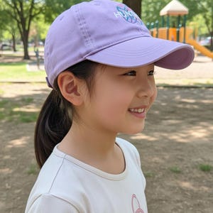 Puede incluir: Gorra de b&eacute;isbol lila con visera curva. La gorra tiene un logotipo y la lleva una persona. La persona lleva una camiseta blanca y sonr&iacute;e. El fondo es un parque con &aacute;rboles y un parque infantil.