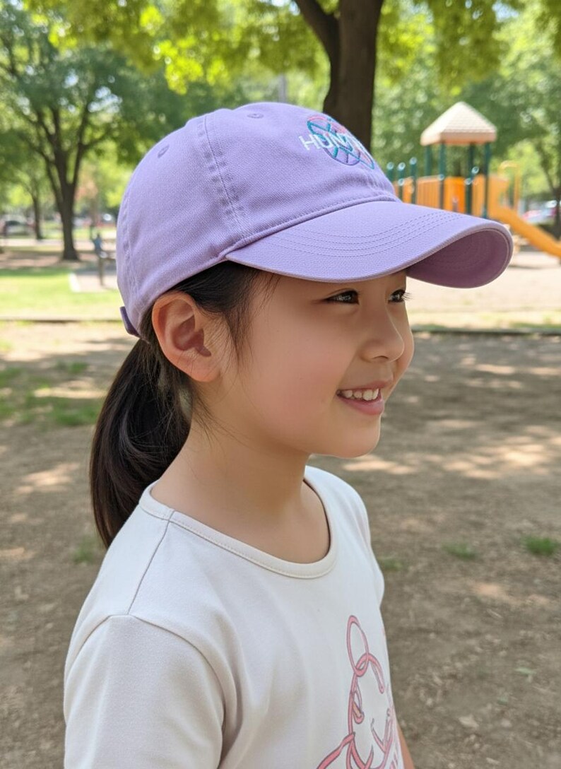 Puede incluir: Gorra de b&eacute;isbol lila con visera curva. La gorra tiene un logotipo y la lleva una persona. La persona lleva una camiseta blanca y sonr&iacute;e. El fondo es un parque con &aacute;rboles y un parque infantil.