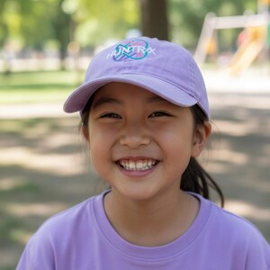 Puede incluir: Una persona con una gorra de b&eacute;isbol lavanda con la palabra "HUNTRAX" en un dise&ntilde;o colorido. La persona tambi&eacute;n lleva una camiseta lavanda. El fondo es un parque con &aacute;rboles y juegos.