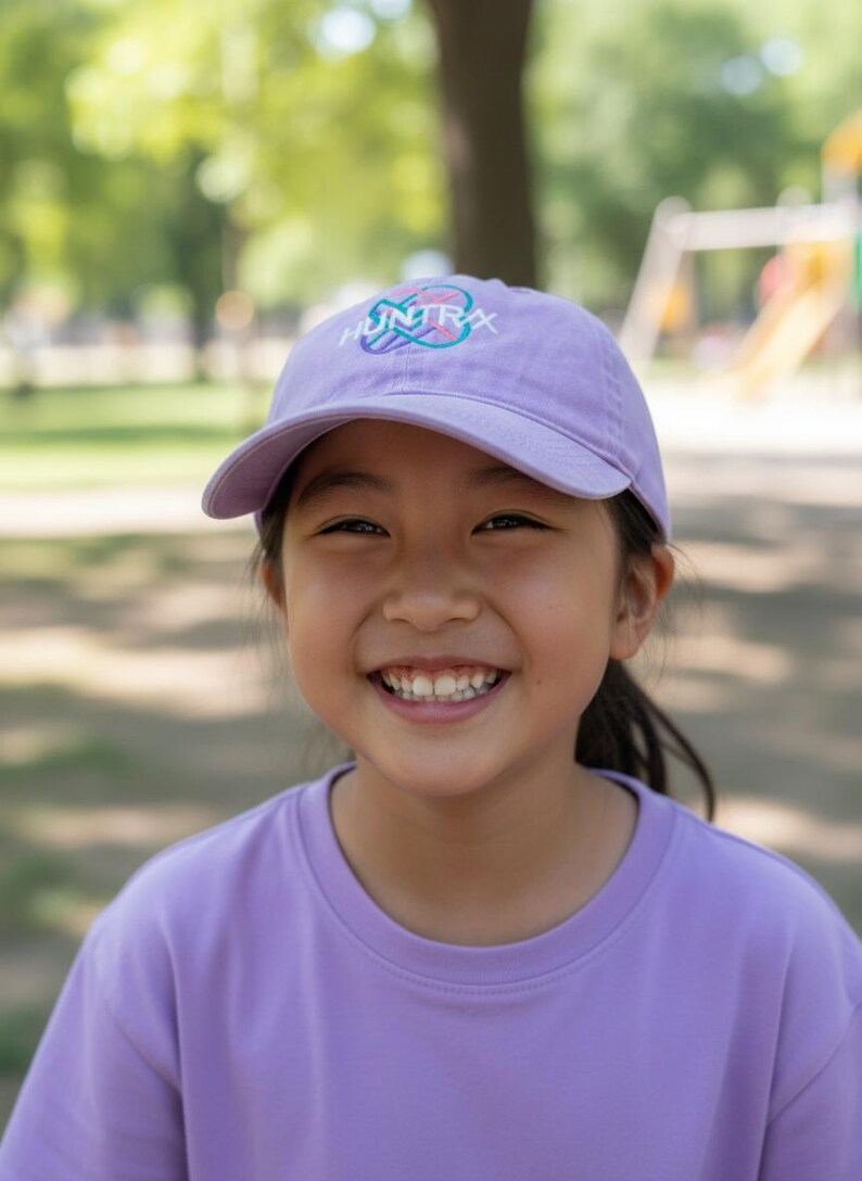 Puede incluir: Una persona con una gorra de b&eacute;isbol lavanda con la palabra "HUNTRAX" en un dise&ntilde;o colorido. La persona tambi&eacute;n lleva una camiseta lavanda. El fondo es un parque con &aacute;rboles y juegos.