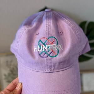 Puede incluir: Gorra de b&eacute;isbol lavanda con la palabra "HUNTRAX" bordada en blanco, con un dise&ntilde;o geom&eacute;trico colorido. La gorra tiene una visera curva y un bot&oacute;n en la parte superior. Hecha de tela suave.