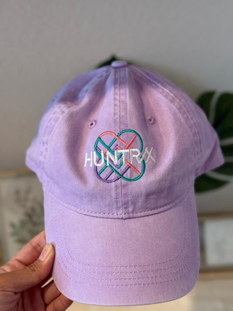 Puede incluir: Gorra de b&eacute;isbol lavanda con la palabra "HUNTRAX" bordada en blanco, con un dise&ntilde;o geom&eacute;trico colorido. La gorra tiene una visera curva y un bot&oacute;n en la parte superior. Hecha de tela suave.