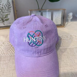 Puede incluir: Gorra de b&eacute;isbol lavanda con la palabra "HUNTRX" bordada en blanco, turquesa, rosa y morado. La gorra tiene una visera curva y un bot&oacute;n en la parte superior. El sombrero est&aacute; hecho de tela suave.