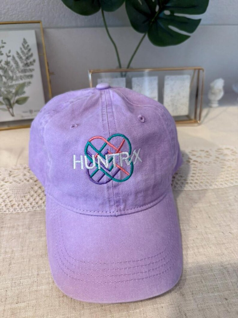 Puede incluir: Gorra de b&eacute;isbol lavanda con la palabra "HUNTRX" bordada en blanco, turquesa, rosa y morado. La gorra tiene una visera curva y un bot&oacute;n en la parte superior. El sombrero est&aacute; hecho de tela suave.