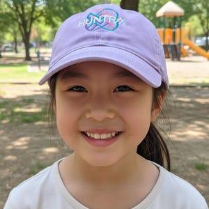 Puede incluir: Una persona joven con una gorra de b&eacute;isbol lavanda con la palabra "HUNTRAX" en letras blancas y turquesas. La persona est&aacute; sonriendo, con un parque infantil visible en el fondo.