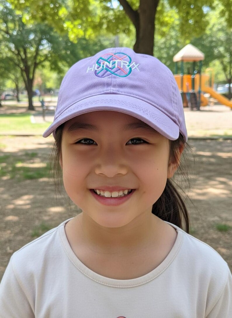 Puede incluir: Una persona joven con una gorra de b&eacute;isbol lavanda con la palabra "HUNTRAX" en letras blancas y turquesas. La persona est&aacute; sonriendo, con un parque infantil visible en el fondo.