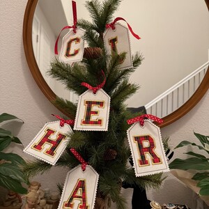 May include: A small Christmas tree decorated with tags spelling out "CHEER". Each tag has a white background with a red and black plaid letter, and is tied with a red ribbon. The tree is in front of a round mirror.
