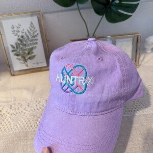 Puede incluir: Gorra de b&eacute;isbol lavanda con la palabra "HUNTRXX" bordada en blanco, turquesa y rosa. La gorra tiene una visera curva y un bot&oacute;n en la parte superior. El sombrero est&aacute; hecho de tela suave.