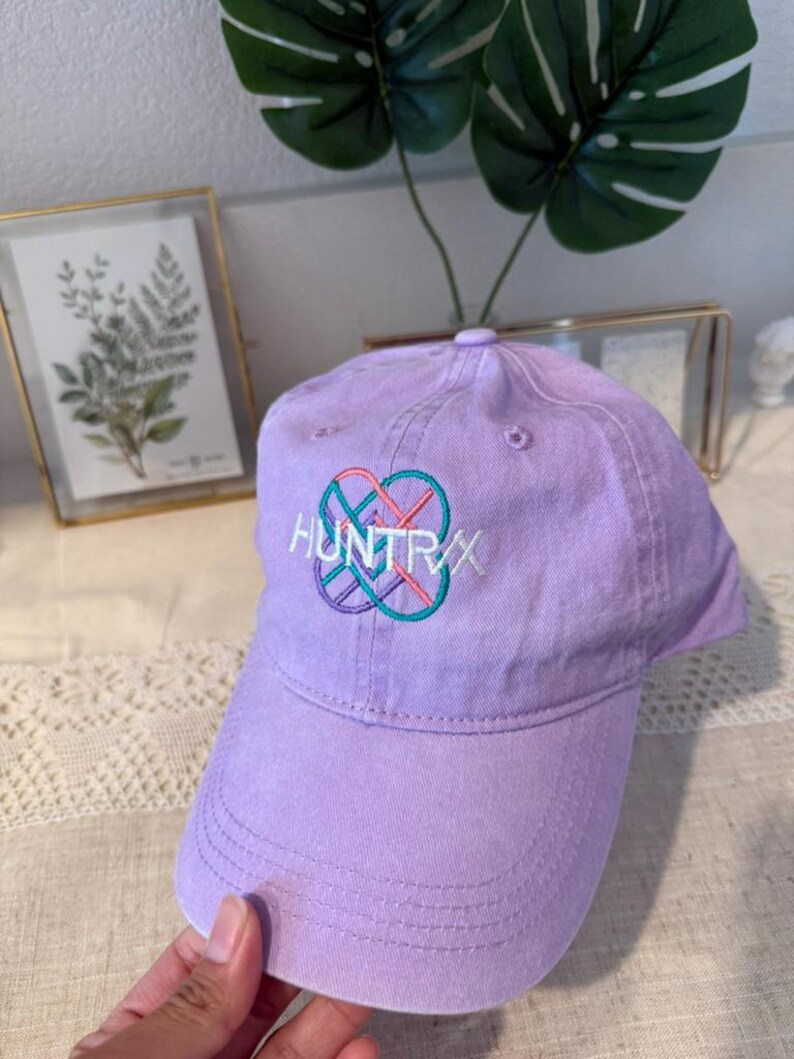 Puede incluir: Gorra de b&eacute;isbol lavanda con la palabra "HUNTRXX" bordada en blanco, turquesa y rosa. La gorra tiene una visera curva y un bot&oacute;n en la parte superior. El sombrero est&aacute; hecho de tela suave.