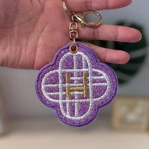 Puede incluir: Un llavero bordado en morado y blanco con un cierre dorado. El llavero tiene forma de trébol de cuatro hojas con un diseño de rejilla blanca y la letra "H" dorada en el centro. Está sujeto a un llavero dorado.