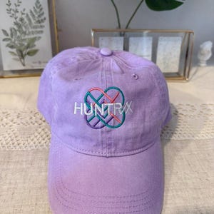 Puede incluir: Gorra de b&eacute;isbol lila con la palabra "HUNTRX" bordada en blanco, turquesa, rosa y morado. La gorra tiene una visera curva y un bot&oacute;n en la parte superior. El sombrero est&aacute; hecho de tela suave.