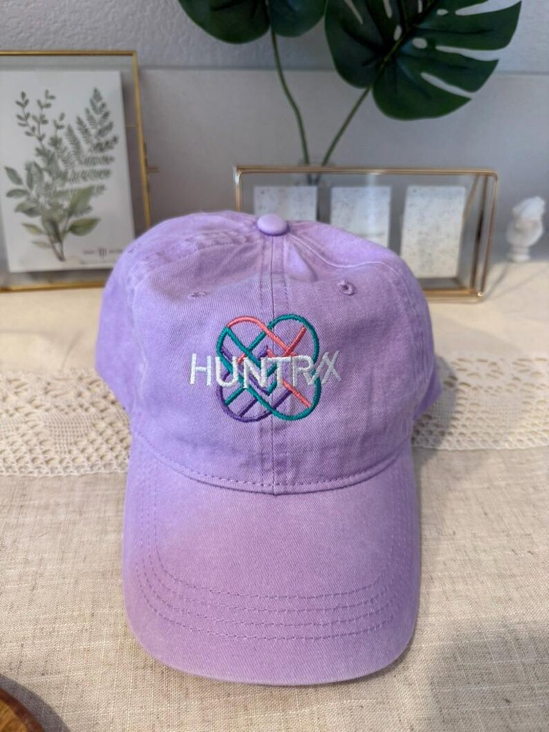 Puede incluir: Gorra de b&eacute;isbol lila con la palabra "HUNTRX" bordada en blanco, turquesa, rosa y morado. La gorra tiene una visera curva y un bot&oacute;n en la parte superior. El sombrero est&aacute; hecho de tela suave.