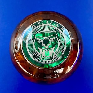 Op de afbeelding: Een ronde, bruin en groene Jaguar-embleem. Het embleem heeft een groene jaguar-kop in het midden, omringd door het woord "JAGUAR" in zilveren letters. De achtergrond heeft een glanzende houtnerf.