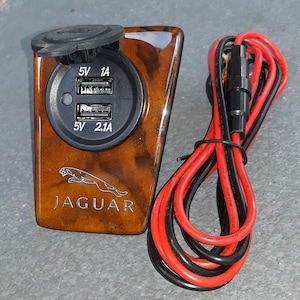 Op de afbeelding: Een auto USB-oplader met een houtnerf afwerking. Het heeft twee USB-poorten met de aanduiding 5V 1A en 5V 2.1A, en het Jaguar-logo. Rode en zwarte draden zijn bevestigd. De oplader heeft een beschermkap.