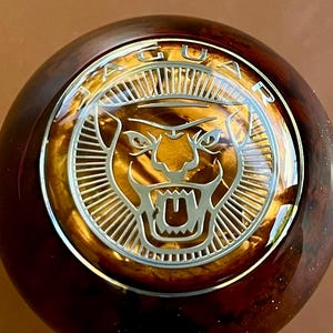 May include: A round gear shift knob featuring a polished dark brown burl wood finish. Centered on the top is a circular silver emblem with the word JAGUAR arched at the top and a stylized silver jaguar head logo set against a golden background.