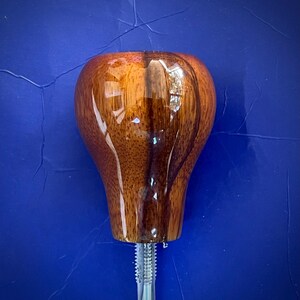 May include: A polished, teardrop-shaped wooden gear shift knob featuring a rich, warm brown wood grain pattern with a glossy finish. The knob is attached to a silver-colored metal threaded bolt extending from the bottom center of the object.