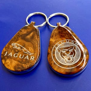May include: Two teardrop-shaped keychains with a polished wood grain finish and silver metal rings. The left keychain features the Jaguar leaping cat logo and text, while the right keychain displays the Jaguar head logo within a circular border.