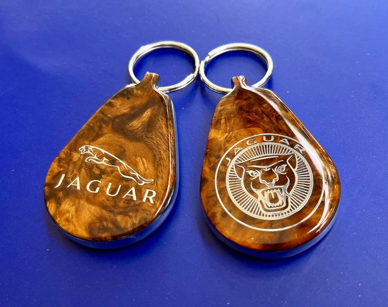 May include: Two teardrop-shaped keychains with a polished wood grain finish and silver metal rings. The left keychain features the Jaguar leaping cat logo and text, while the right keychain displays the Jaguar head logo within a circular border.